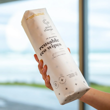 Load image into Gallery viewer, A hand holding a roll of Good Change reusable eco wipes, wrapped in a cream-colored paper sleeve with black text, against a blurred outdoor background.