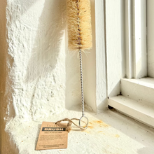 Load image into Gallery viewer, A long-handled cleaning brush with a white fluffy brush head, natural tan bristles, and a silver metal handle, leaning against a sunlit white wall next to a window.