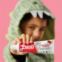 Load image into Gallery viewer, A child wearing a green dinosaur costume holds a tube of Grants of Australia Strawberry Surprise Natural Toothpaste against a solid pink background.