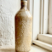 Load image into Gallery viewer, A tall ceramic bottle with a glossy beige and brown floral pattern, featuring a black pour spout, set against a white textured wall and window frame.
