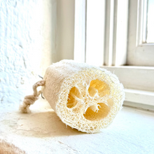 Load image into Gallery viewer, A natural loofah sponge bath brush with a twisted rope handle, resting on a white windowsill in bright natural light.