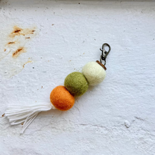 Load image into Gallery viewer, A handmade felt keychain charm with three felt balls in orange, olive green, and cream, finished with a white tassel and a silver metal clasp, resting on a textured white surface.