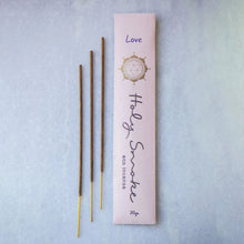 Load image into Gallery viewer, Three incense sticks next to their light pink packaging box on a textured light blue background. The box features a gold geometric emblem, the word 'Love' in purple, and 'Holy Smoke' in cursive script.
