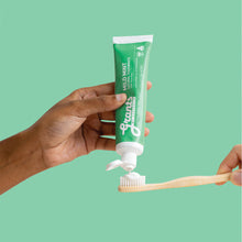 Load image into Gallery viewer, Hand applying St. Tropez toothpaste to a wooden toothbrush against a green background
