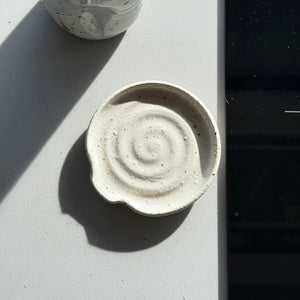 Ceramic bowl with spiral design on a light surface next to a speckled ceramic bottle and a black electronic device.
