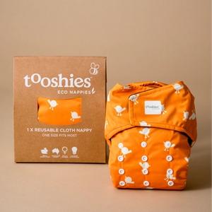 Orange reusable cloth nappy with 'tooshies' branding next to its packaging on a beige background