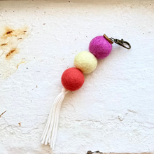 Load image into Gallery viewer, A handmade felted wool keychain with three colorful felt balls in pink, yellow, and red, finished with a white tassel and a metal lobster clasp, resting on a textured white surface.
