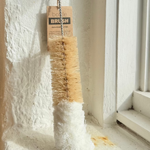 Load image into Gallery viewer, A bottle brush with natural tan bristles and a white soft handle, hanging from a metal hook on a textured white wall next to a window.
