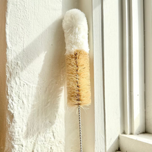 Load image into Gallery viewer, A long-handled cleaning brush with a white fluffy brush head, natural tan bristles, and a silver metal handle, leaning against a sunlit white wall next to a window.
