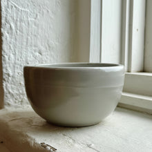 Load image into Gallery viewer, A small round ceramic bowl with a matte, light gray finish sits on a weathered stone windowsill next to a white window frame.
