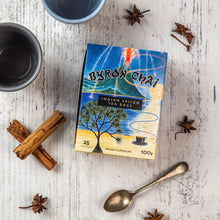 Load image into Gallery viewer, A box of Byron Chai Indian Spiced Tea Bags sits on a white rustic wooden surface, surrounded by tea-related props including cinnamon sticks, star anise, a metal cup, a spoon, and a small dish.
