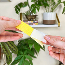 Load image into Gallery viewer, Two hands holding a yellow and white tape with a plant and shelf in the background
