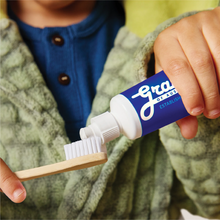 Load image into Gallery viewer, A close-up of a child's hands applying toothpaste from a white and blue tube onto a small wooden toothbrush.
