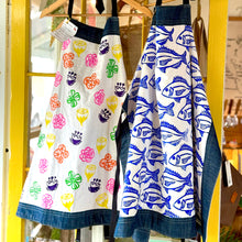 Load image into Gallery viewer, Two aprons with colorful and fish patterns hanging on a yellow frame.
