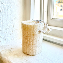 Load image into Gallery viewer, A natural beige loofah sponge with rope handles, standing on a white windowsill next to a textured white wall.
