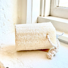 Load image into Gallery viewer, A rectangular natural sponge washcloth with a knotted cord handle, resting on a sunlit windowsill.
