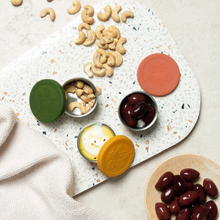 Load image into Gallery viewer, Nuts and olives in containers on a speckled stone board with colorful lids.

