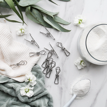 Load image into Gallery viewer, A styled flat-lay of several silver metal spring clips arranged on a white marble surface with a jar of white powder, a spoonful of powder, a textured cream towel, a sage green towel, white flowers, and eucalyptus leaves.
