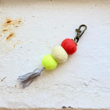 Load image into Gallery viewer, A handmade felt keychain with three colorful felt balls in red, cream, and lime green, finished with a gray tassel and an antique brass key clasp, resting on a textured off-white surface.
